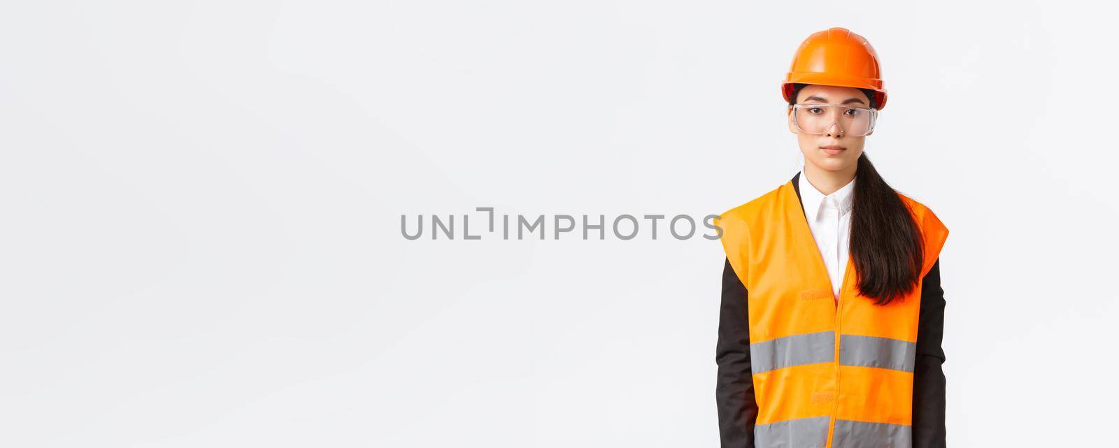 Serious-looking professional female asian construction engineer, architect at building area wearing uniform, safety helmet over business suit, standing white background, inspect enterprise.