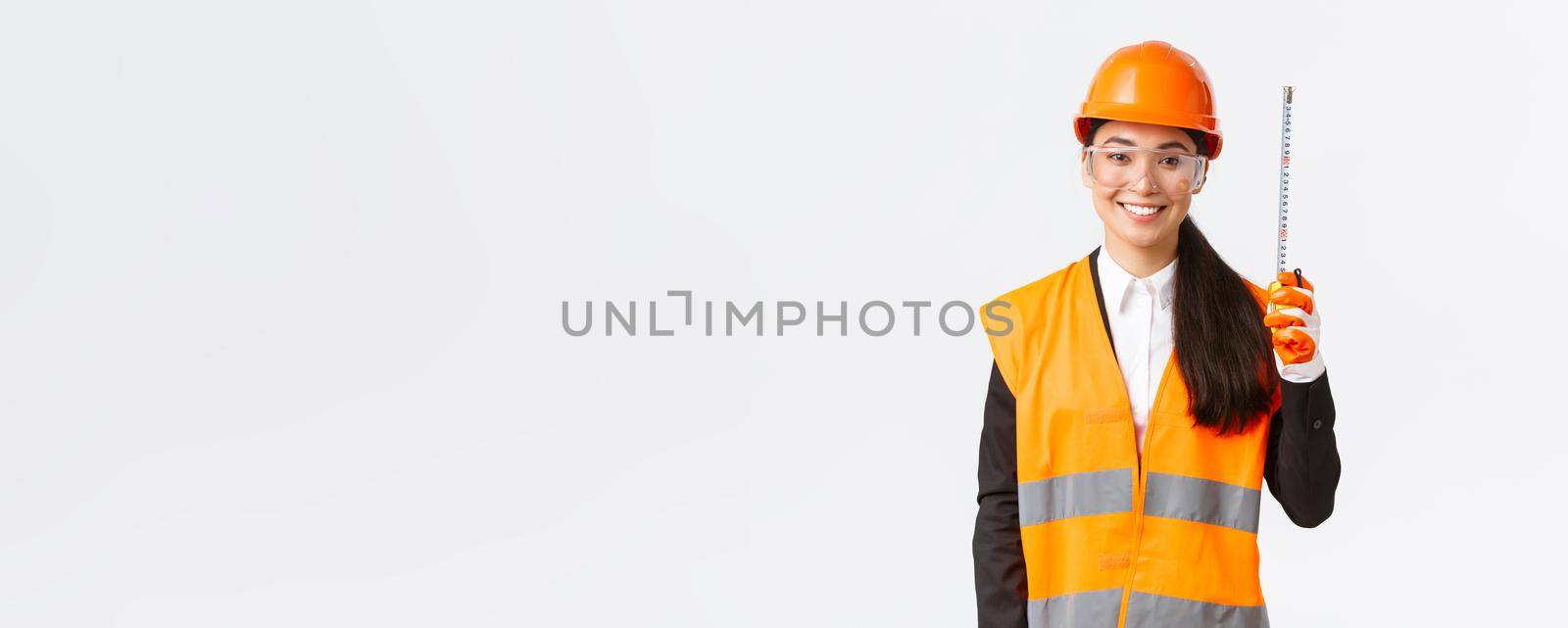 Confident smiling, professional asian female engineer, construction technician in safety helmet and reflective uniform, standing with tape measure, measuring layout at building area.
