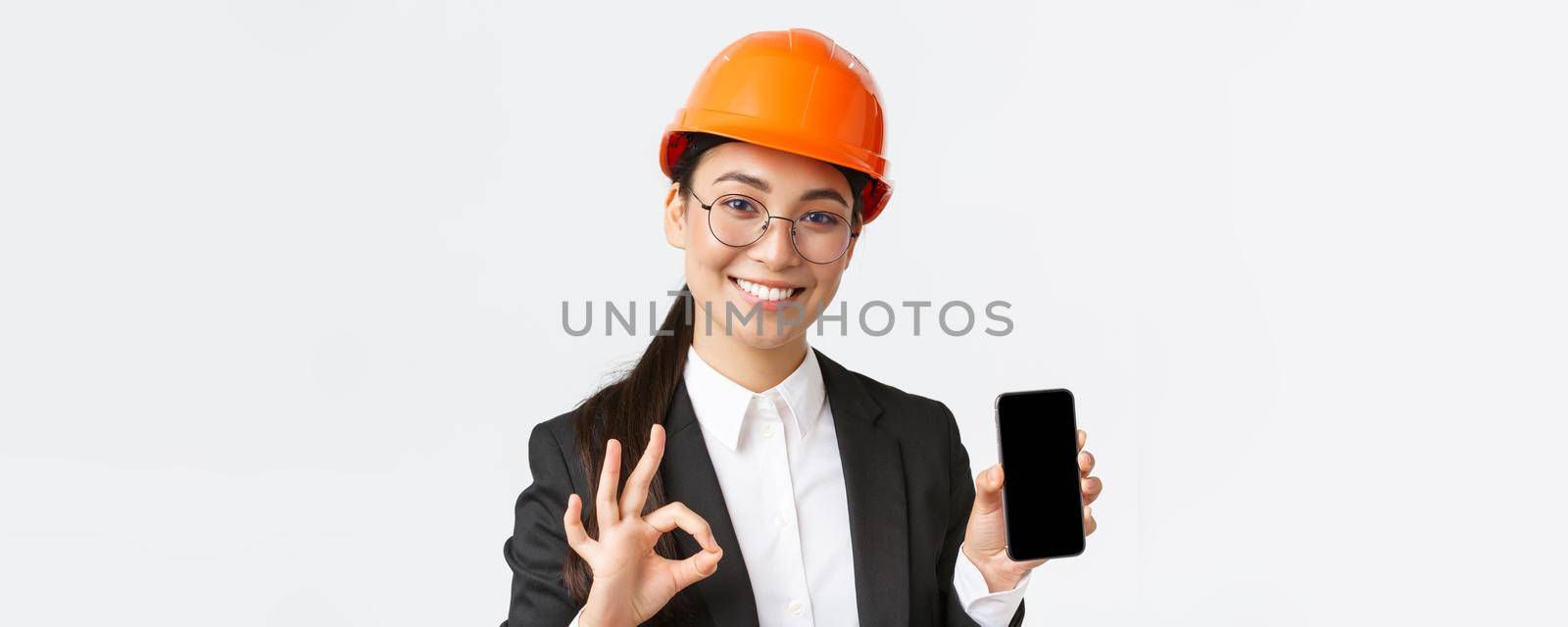 Satisfied young female asian engineer, architect in suit and safety helmet showing mobile phone screen and make okay gesture, approve, recommend application, white background.