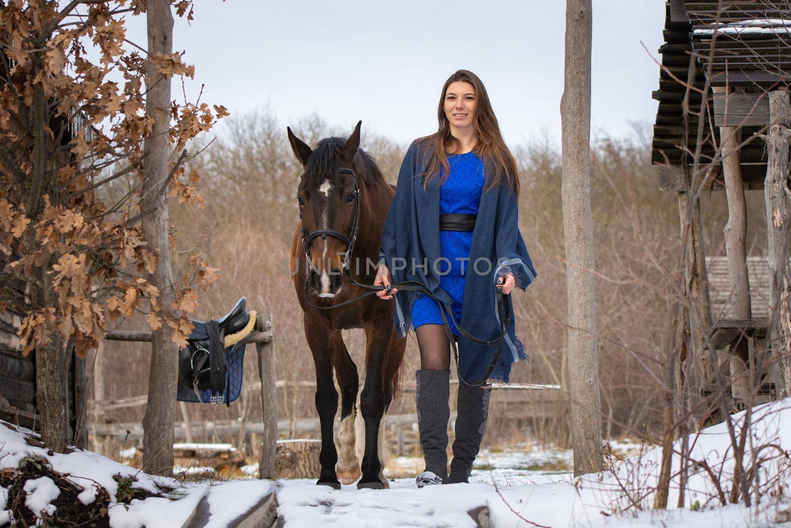 A girl in a blue dress walks with a horse through the winter forest and old wooden ruins