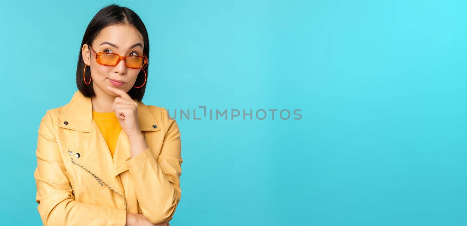 Portrait of asian woman thinking, looking thoughtful, searching ideas or solution, wearing sunglasses, standing over blue background.