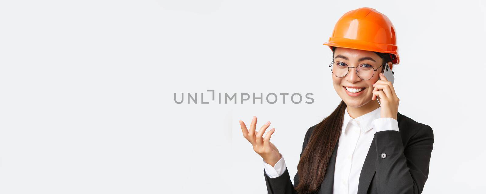 Close-up of asian businesswoman manage enterprise, engineer in safety helmet and suit having phone conversation, calling investors, smiling while talking over smartphone, white background by Benzoix