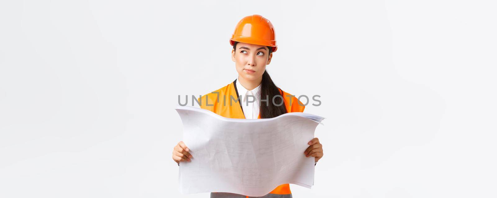 Thoughtful professional asian female construction manager, architect reading blueprints and looking away pondering, thinking, making decision while studying project plan, white background.