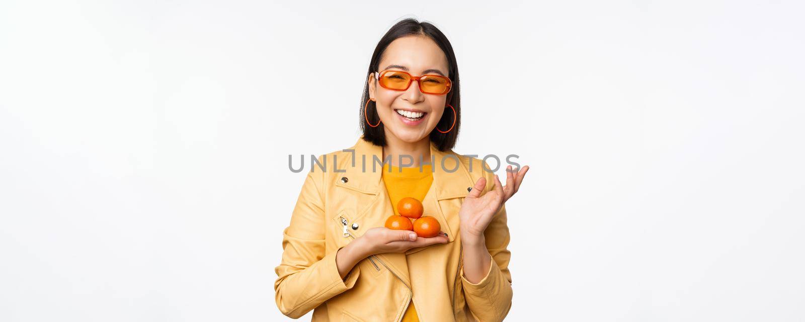 Stylish happy asian girl in sunglasses holding tangerines and smiling, posing against white background by Benzoix
