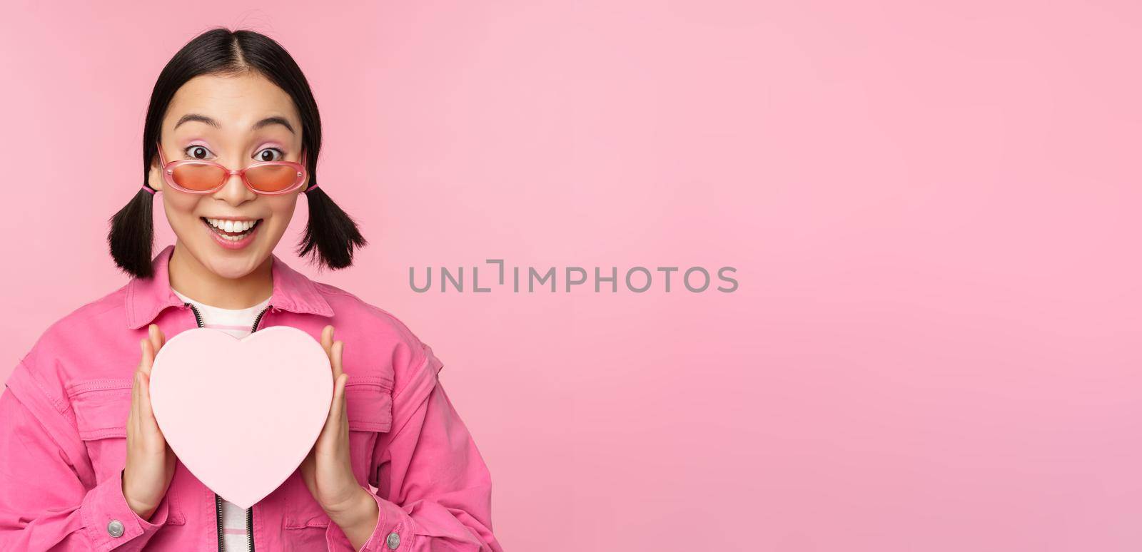 Beautiful asian girl smiling happy, showing heart gift box and looking excited at camera, standing over pink romantic background.