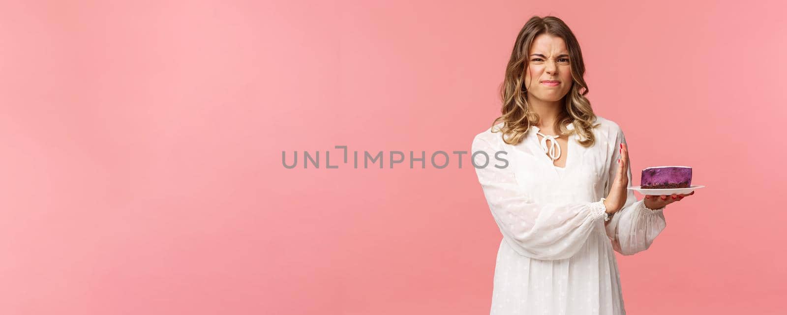 Holidays, spring and party concept. Portrait of dissatisfied and displeased blond girl in white dress rejecting, declining eat tasteless cake, grimacing and show stop sign at dessert, pink background by Benzoix