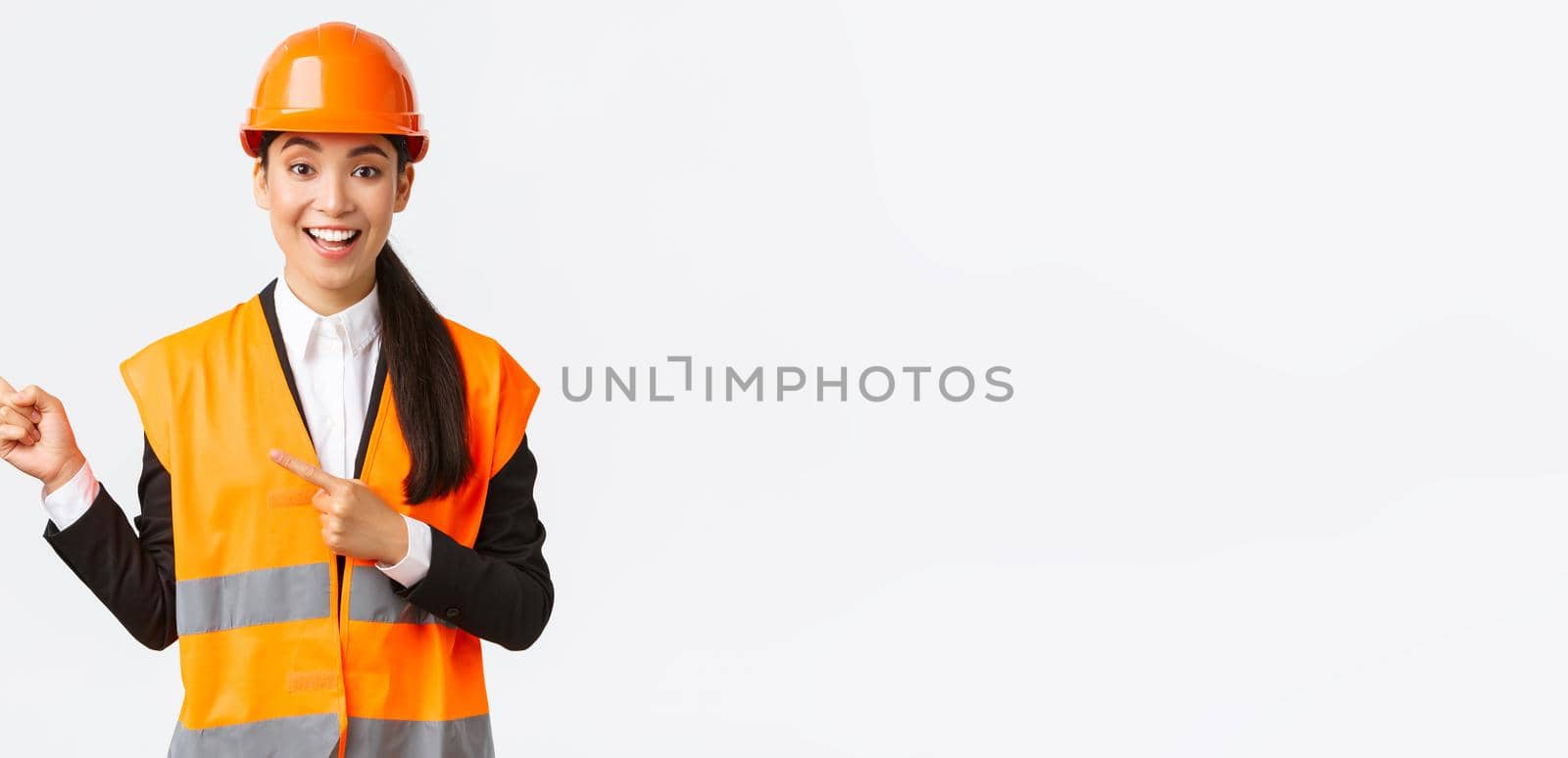 Building, construction and industrial concept. Smiling female engineer in helmet and reflective clothing showing way, pointing upper left corner, talking about constructing project, white background by Benzoix