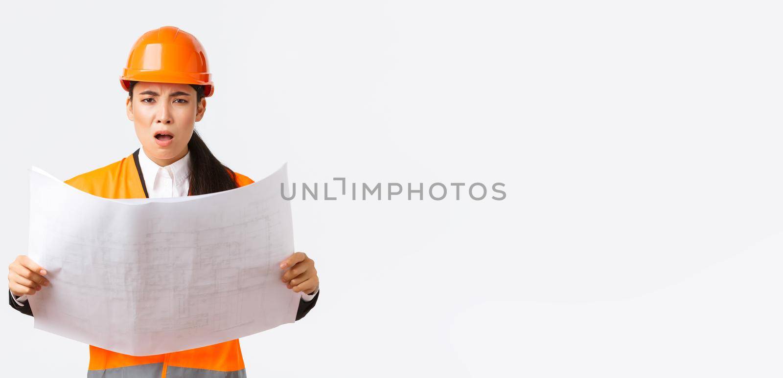 Frustrated and displeased female asian engineer, chief architect at construction zone, looking disappointed after studying blueprints, stare camera, scolding manager, white background.