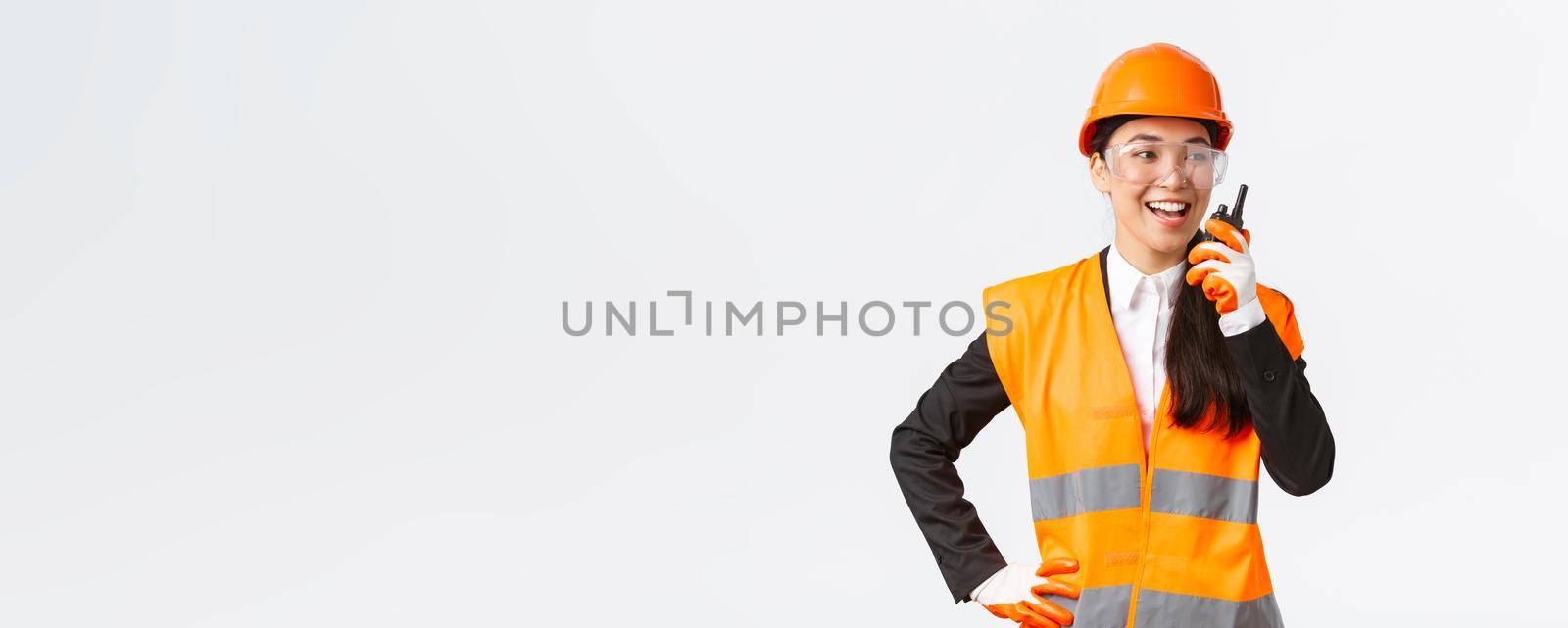 Confident pleased asian female engineer in safety helmet and uniform talking with chief architect using walkie-talkie. Satisfied construction technician contact team using radio phone by Benzoix