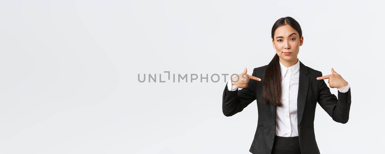 Professional asian businesswoman in suit pointing at herself as offering best deals. Proud and confident saleswoman show-off, suggest personal help or assistance, standing white background.