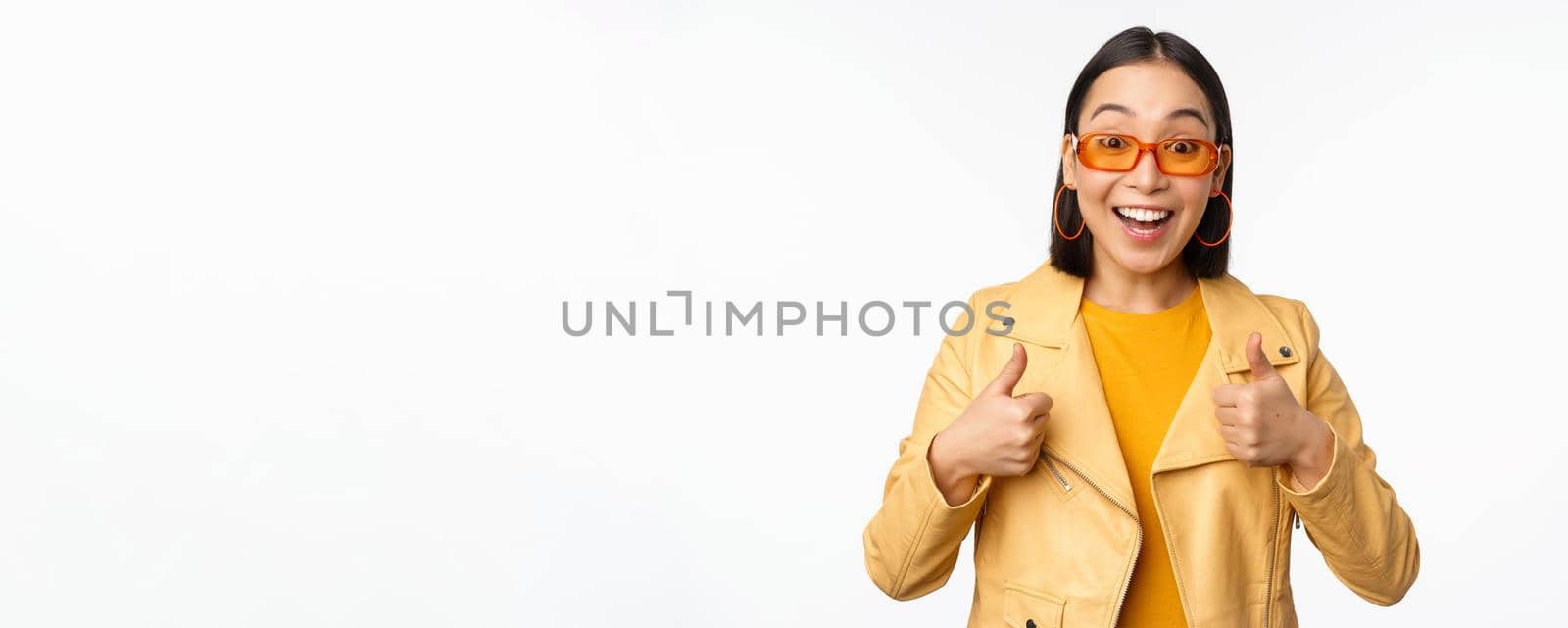 Enthusiastic asian girl smiles, shows thumbs up and looks happy, saying yes, approve and recommend smth, standing over white background.