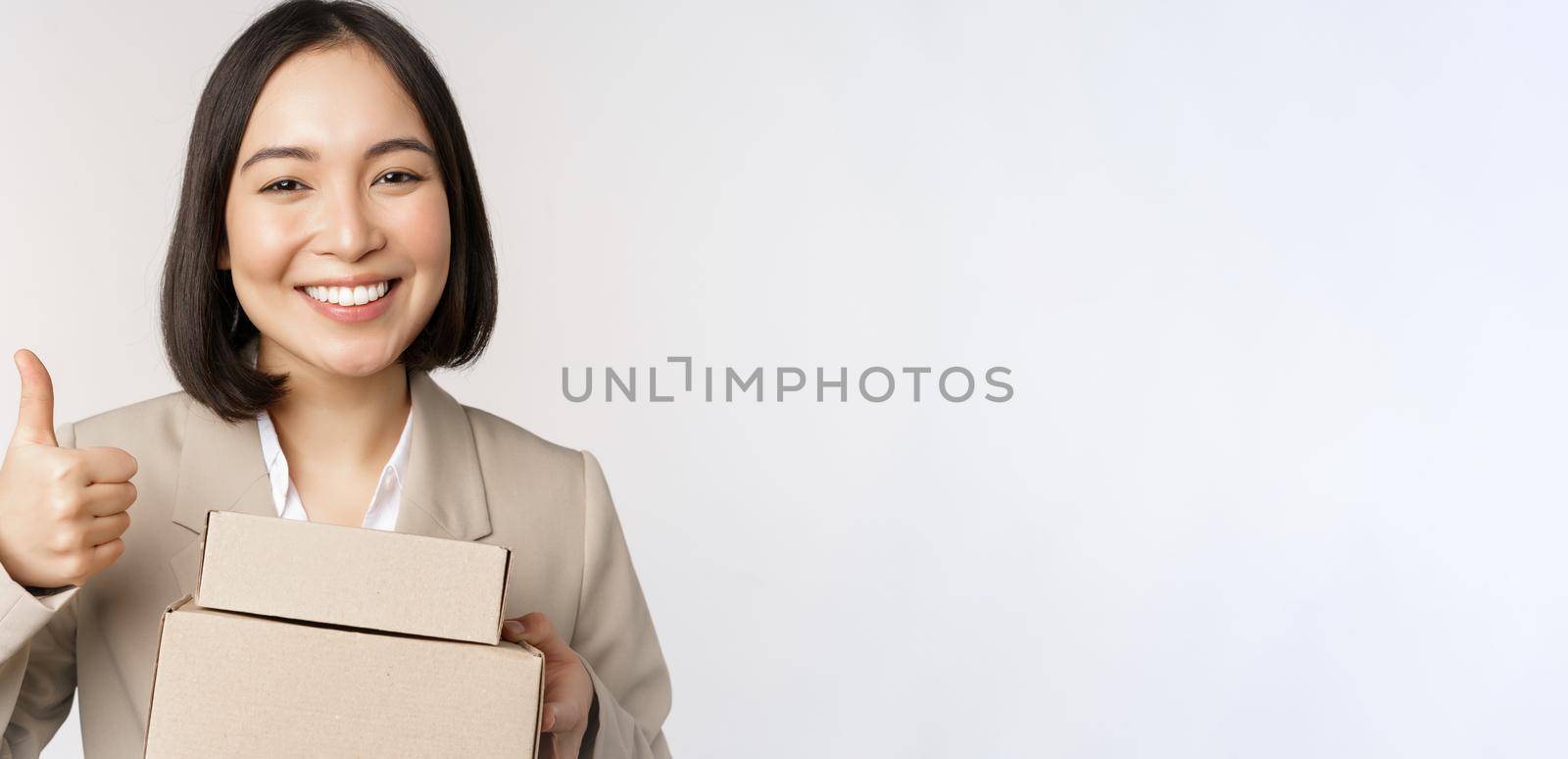 Smiling asian businesswoman, showing thumbs up and boxes with delivery goods, prepare order for client, standing over white background.