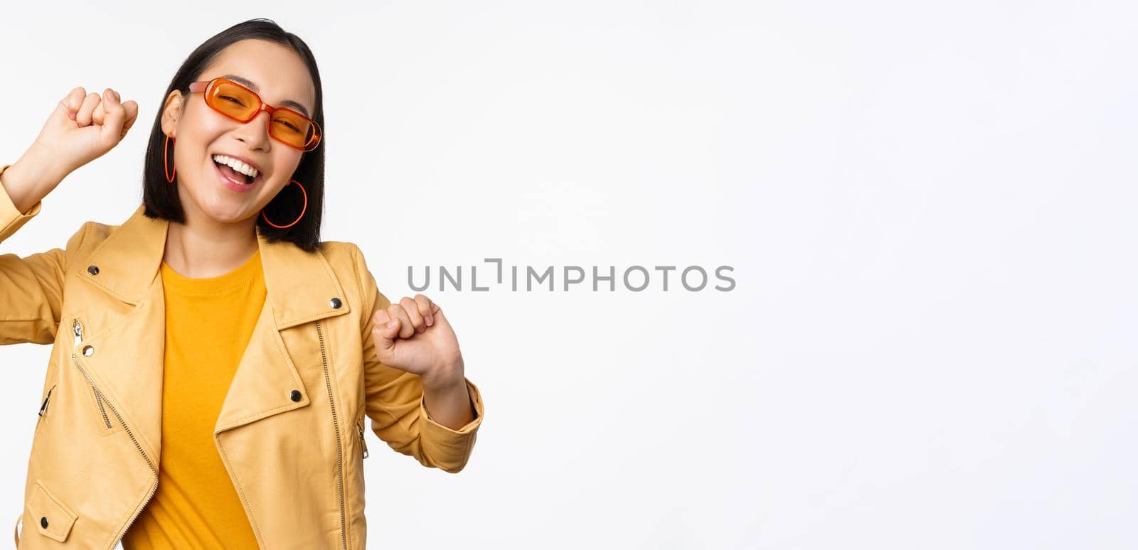 Happy stylish korean girl in sunglasses, dancing and laughing, smiling carefree, standing over white background. Copy space