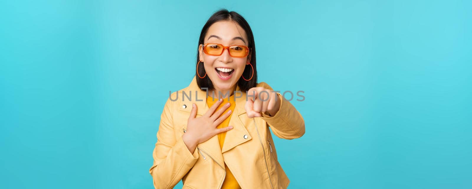 Image of happy korean woman in sunglasses, pointing finger at camera with amazed, surprised and joyful face expression, standing over blue background. Copy space