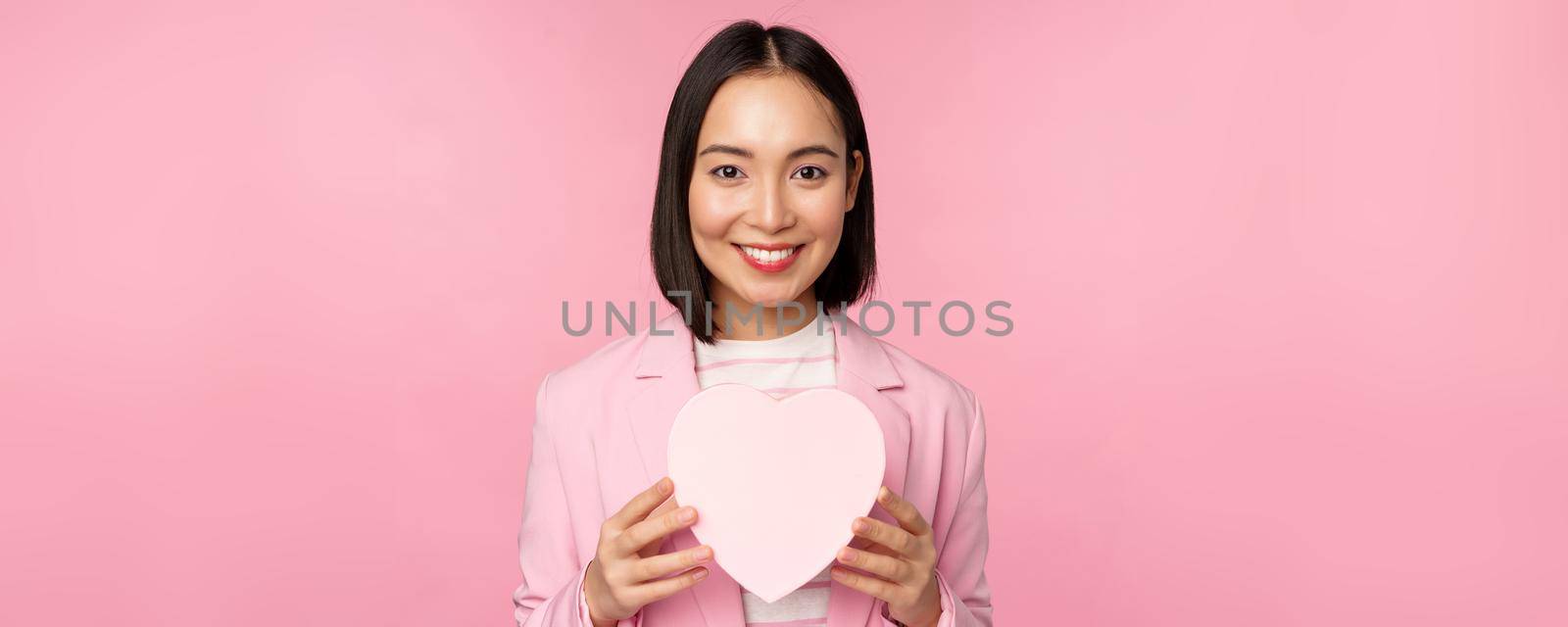 This is for you. Romantic cute asian corporate woman, girl in suit, showing heart-shaped box with gift, standing over pink background by Benzoix