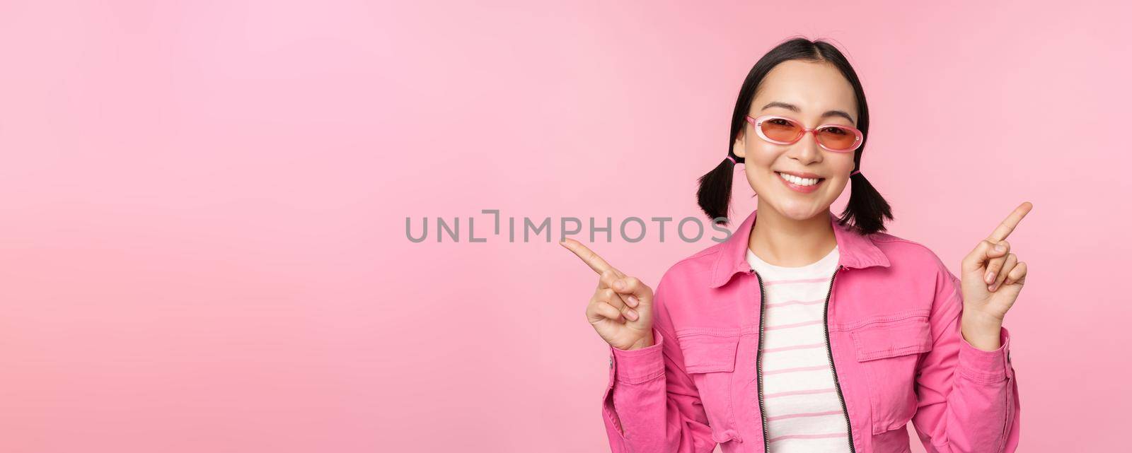 Choice. Stylish korean girl, asian female model points fingers sideways, shows two variants, product advertisement, demonstrating items, standing over pink background.