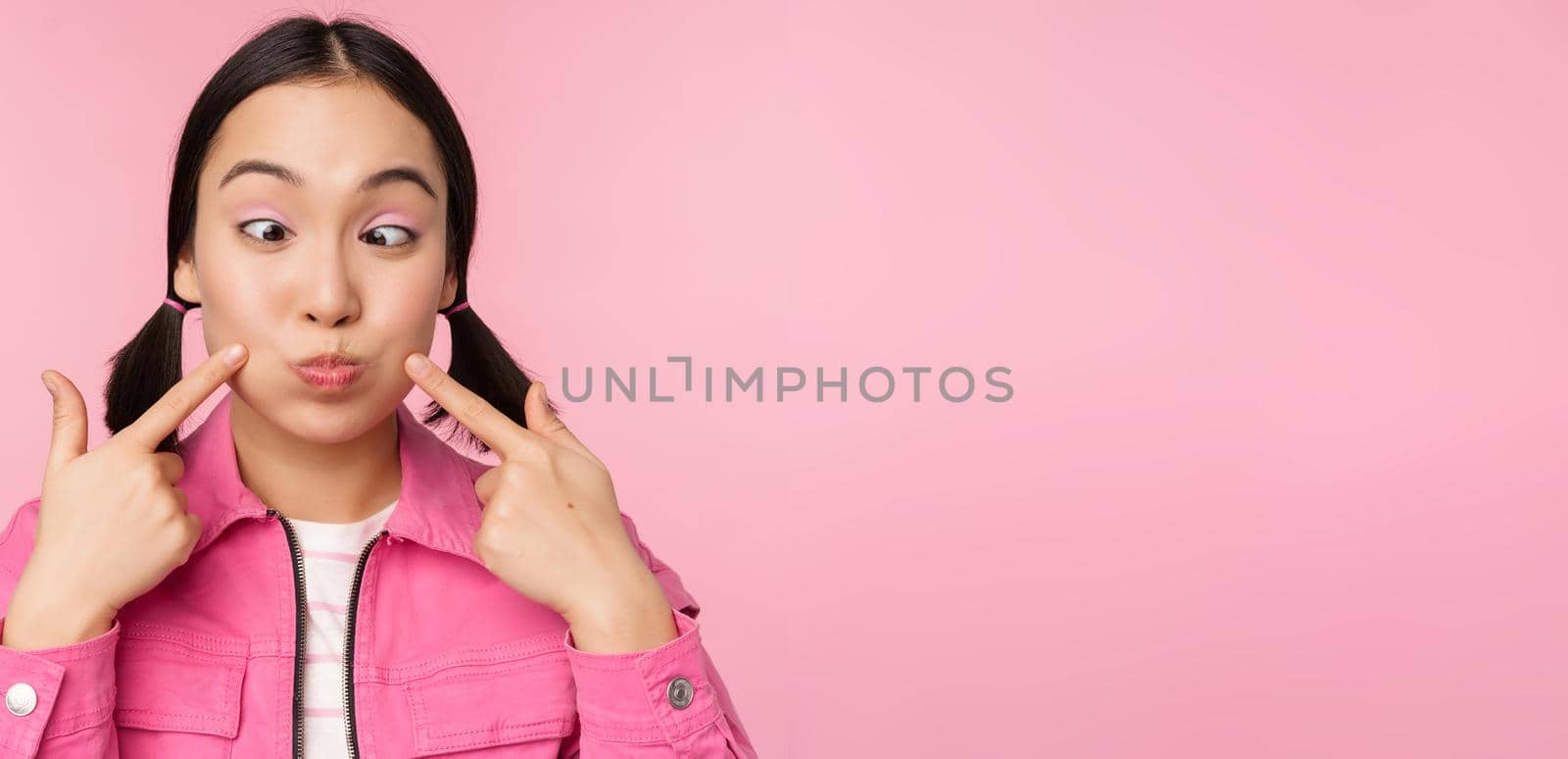 Close up portrait of young asian girl showing her dimples, poking cheeks silly and making funny faces, standing over pink background.