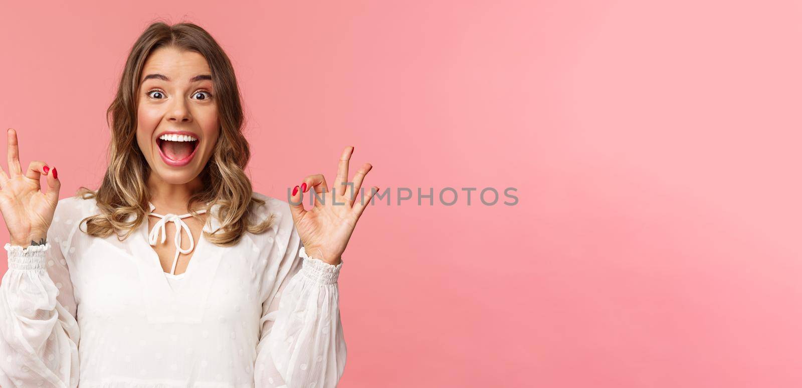 Portrait of excited and upbeat, cheerful blond girl in white dress dancing and showing okay signs with happy beaming smile, excited over music festival, rate awesome movie, recommend product by Benzoix