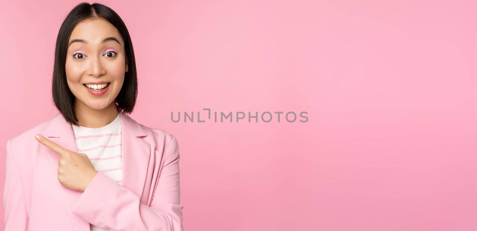 Portrait of asian business woman, saleswoman in suit pointing finger left, showing banner advertisement, smiling and looking professional, pink background.