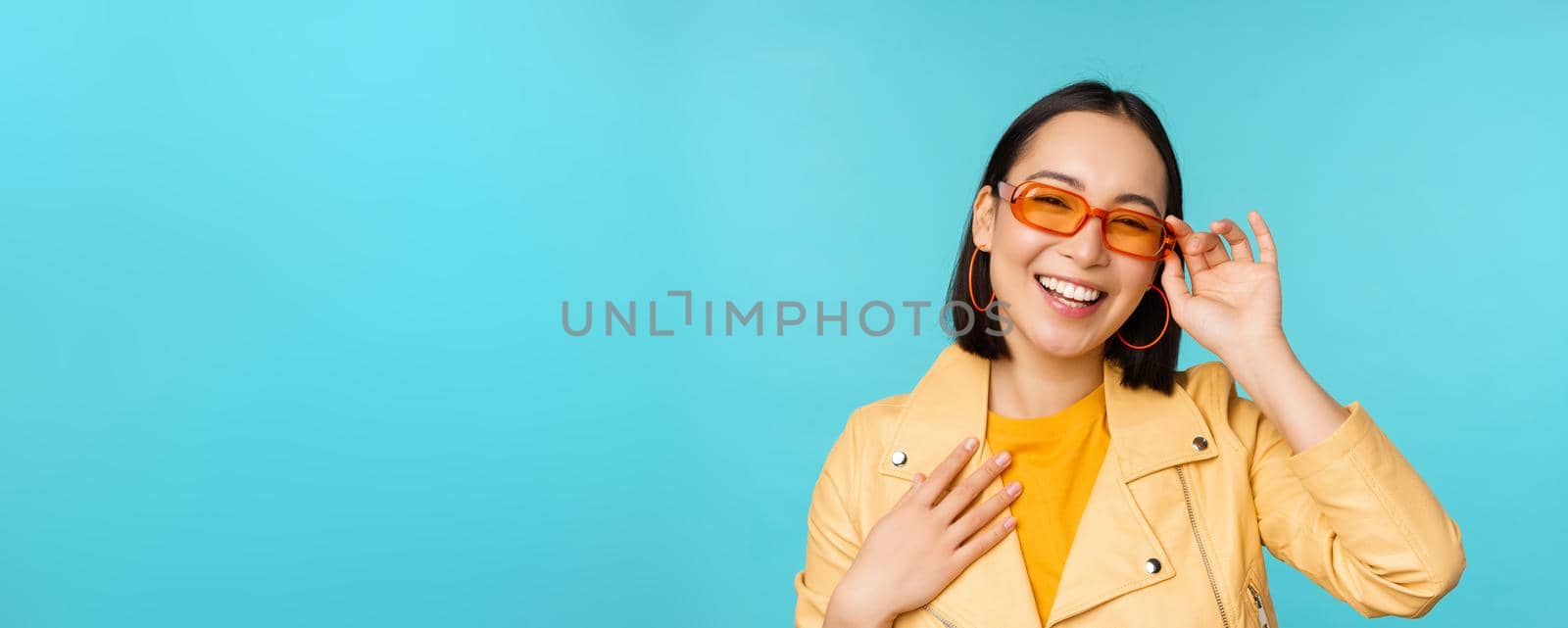 Close up portrait of stylish asian woman in sunglasses, laughing and smiling, looking happy, posing in trendy clothes over blue background.