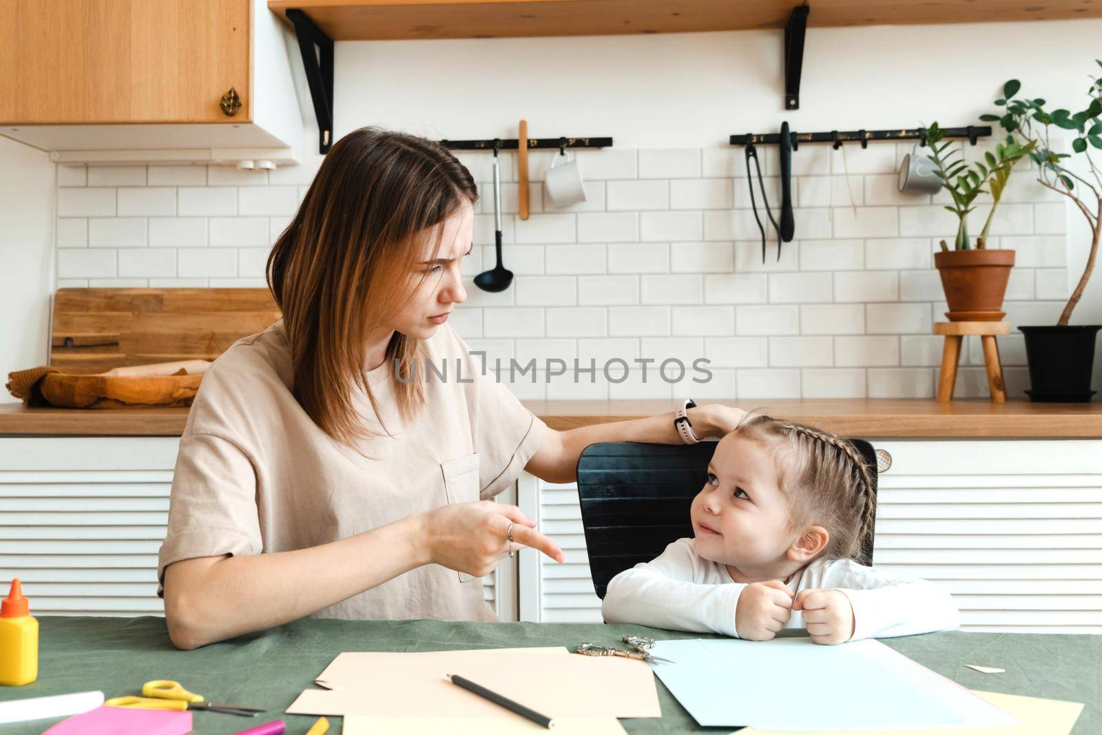Cute angry little preschool girl doing creative homework with her mom while sitting at the table in the living room and arguing. Problems in making crafts for kindergarten