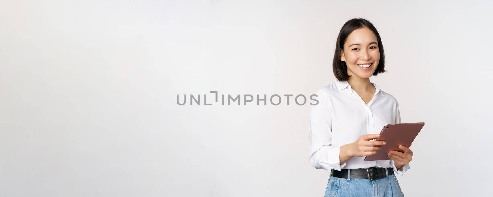 Image of young ceo manager, korean working woman holding tablet and smiling, standing over white background.
