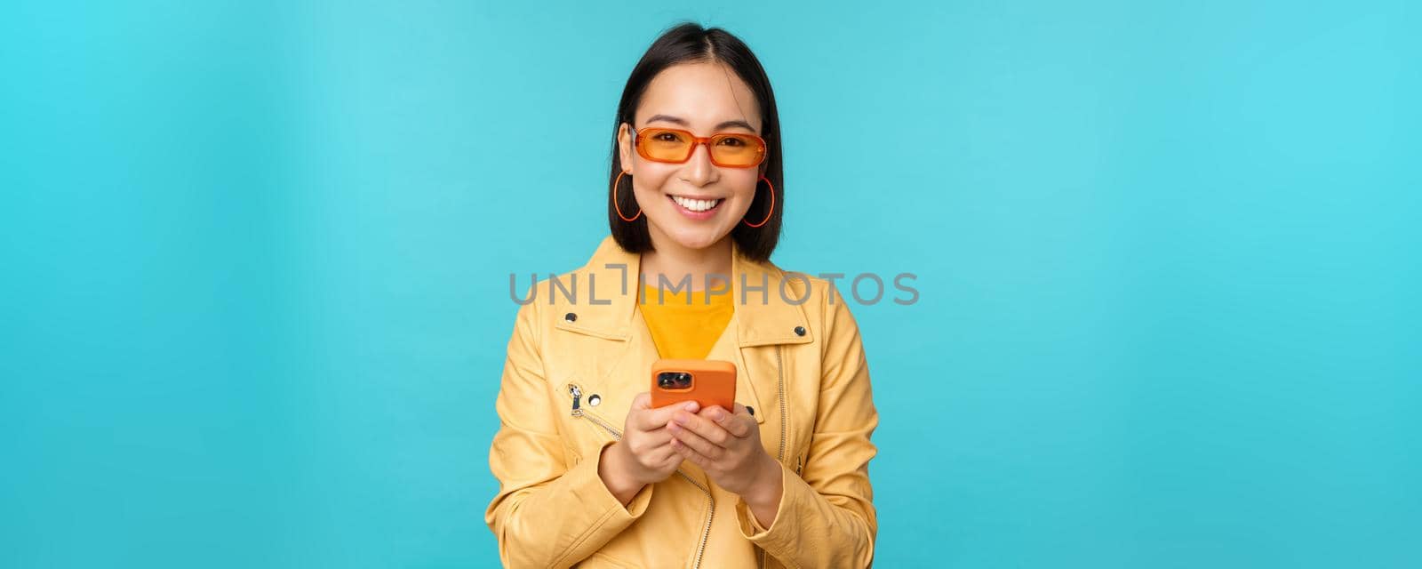 Smiling asian girl in sunglasses, using smartphone app, holding mobile phone, standing over blue background by Benzoix