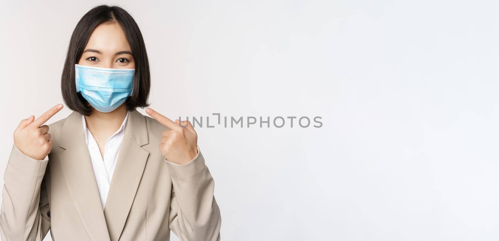 Coronavirus and business people concept. Asian female entrepreneur pointing fingers at medical face mask at workplace, standing over white background.