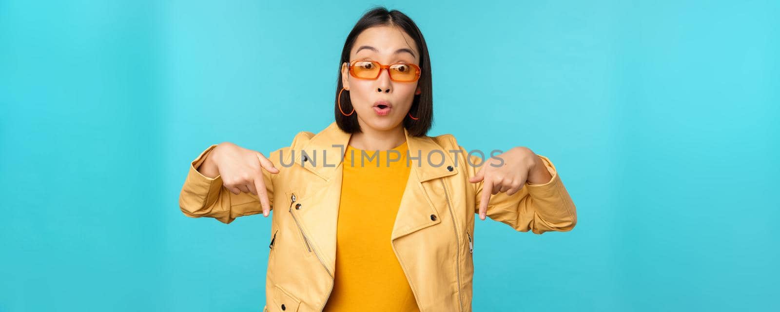 Portrait of stylish asian girl, wears sunglasses, smiles and points fingers down, shows advertisement, stands over blue background.