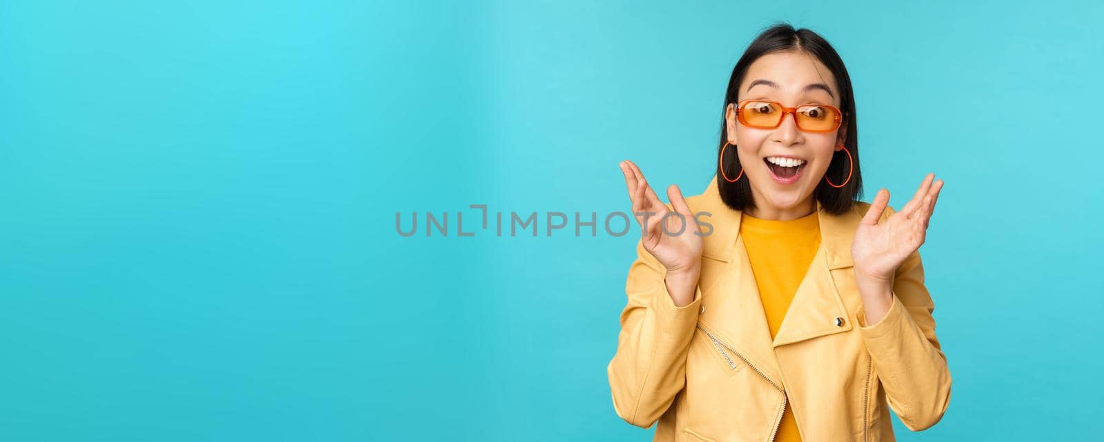 Image of enthusiastic young asian woman celebrating, triumphing, looking surprised and happy, clapping hands satisfied, standing over blue background.