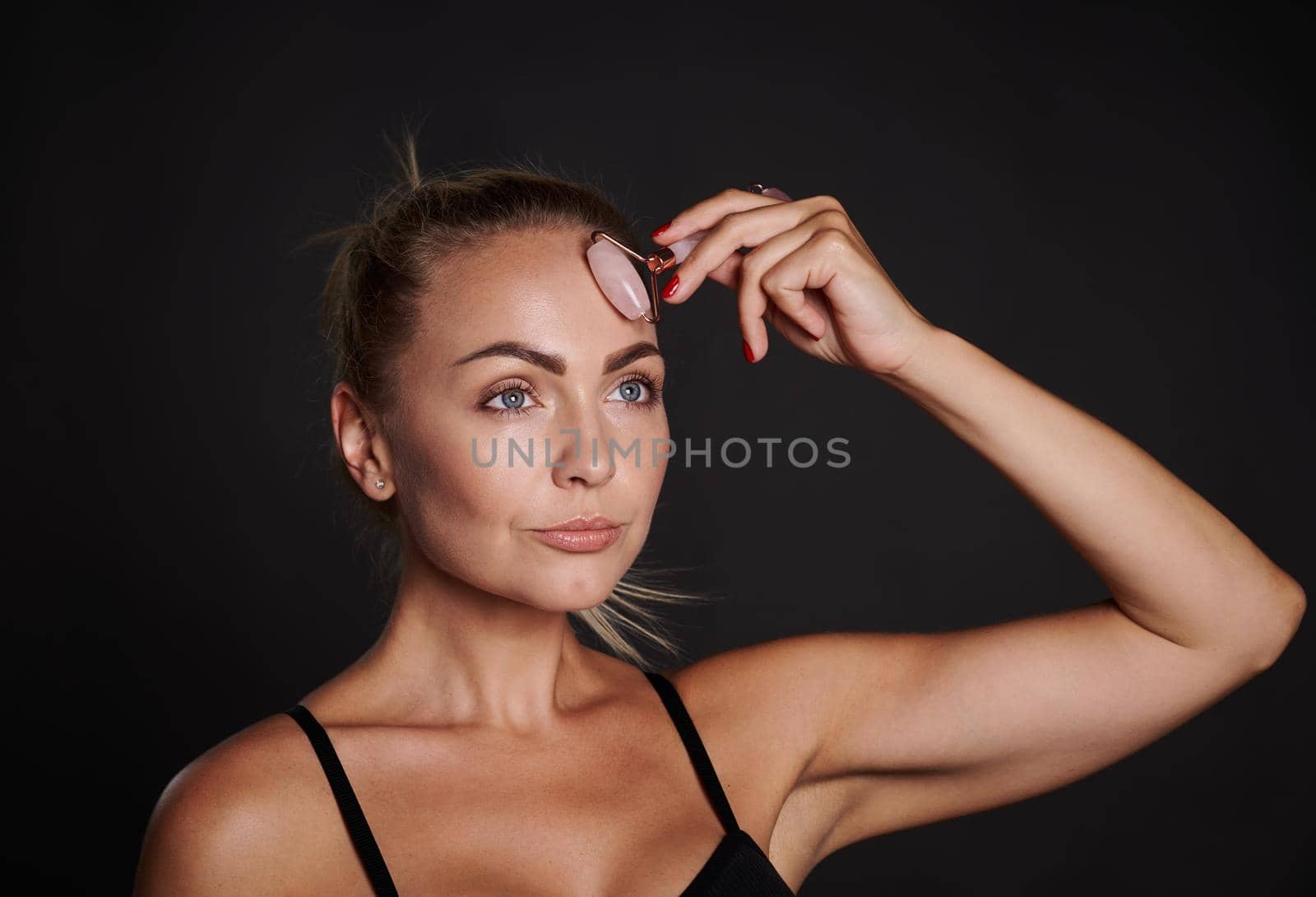 Face portrait of a pretty European woman wearing black underwear doing a lymphatic drainage facial massage with a roller massager, isolated over dark background. Beauty spa treatment, copy space