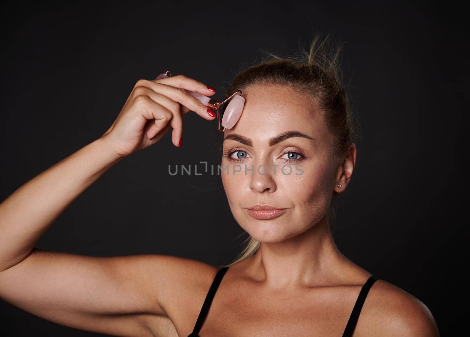 Close up of middle aged Caucasian woman with healthy glowing tanned skin, massaging face with jade roller isolated over black background with copy ad space