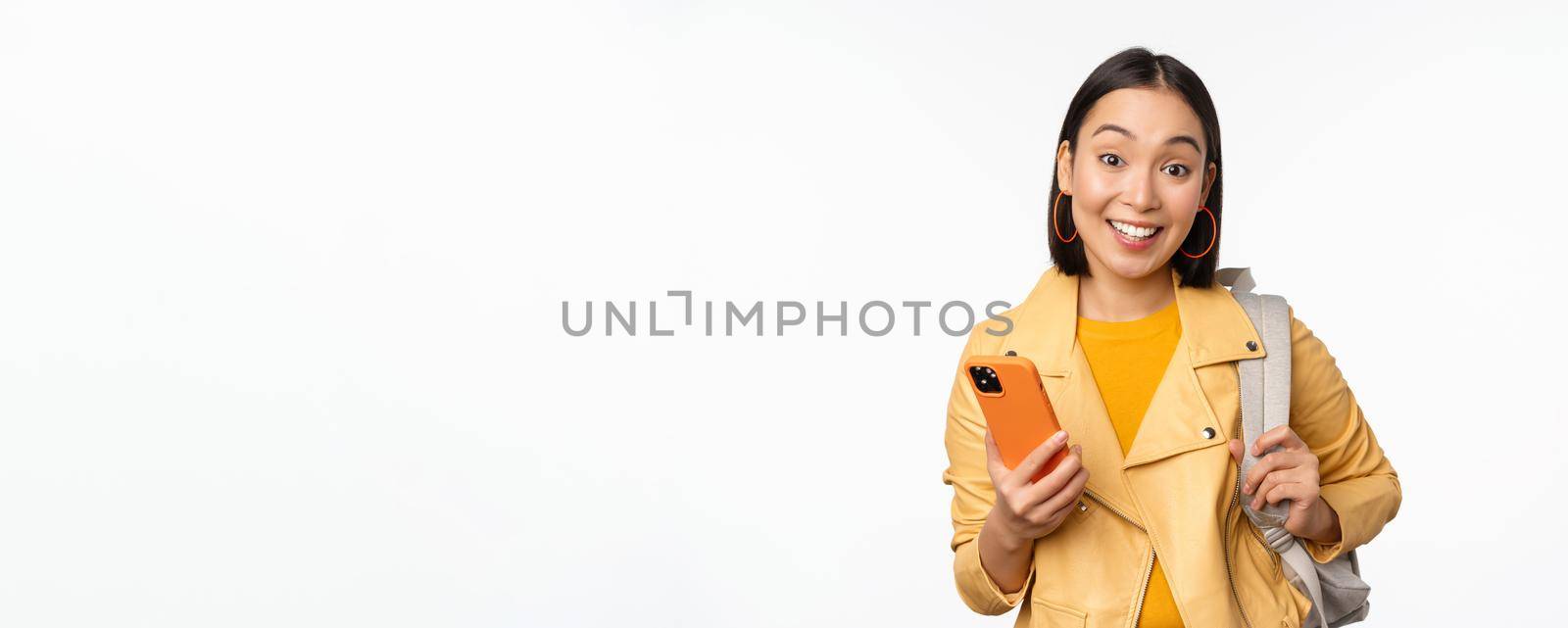 Image of happy girl traveller, tourist with backpack looking at smartphone, using mobile phone route app, standing over white background by Benzoix