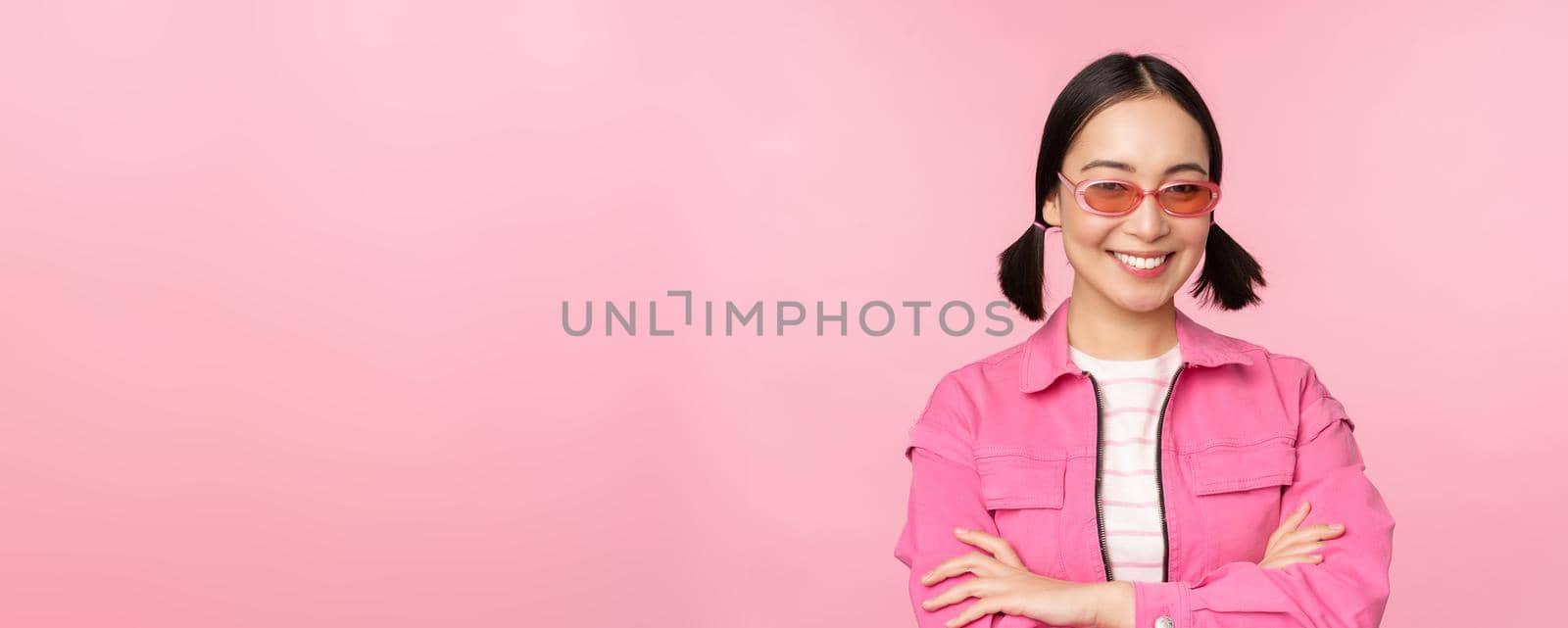 Stylish asian girl in pink clothes and sunglasses, smiling and looking happy at camera, standing over studio background by Benzoix