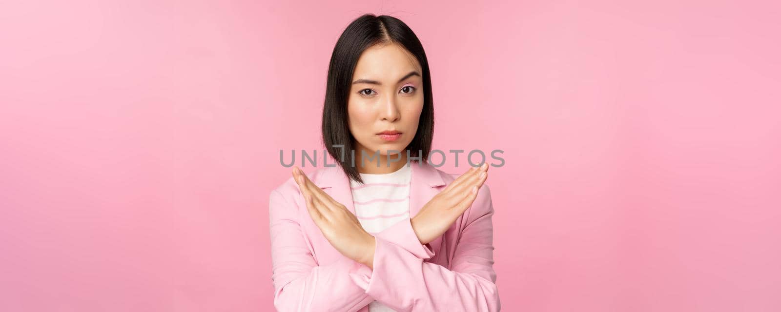 Image of serious corporate woman, asian businesswoman showing stop, cross gesture, disapprove smth, standing in suit over pink background.