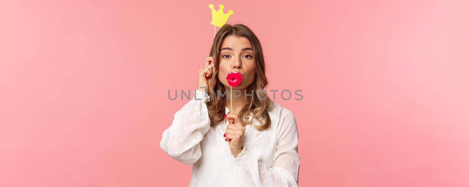 Spring, happiness and celebration concept. Close-up portrait of funny and cute, silly blond girl in white dress, holding big lips mask and crown, partying and having fun, pink background by Benzoix