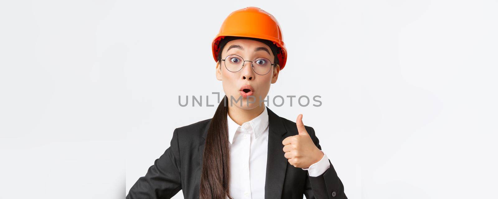 Close-up of professional pleased asian female chief engineer, architect in business suit and safety helmet showing thumbs-up in approval, give permission, recommend construction company.