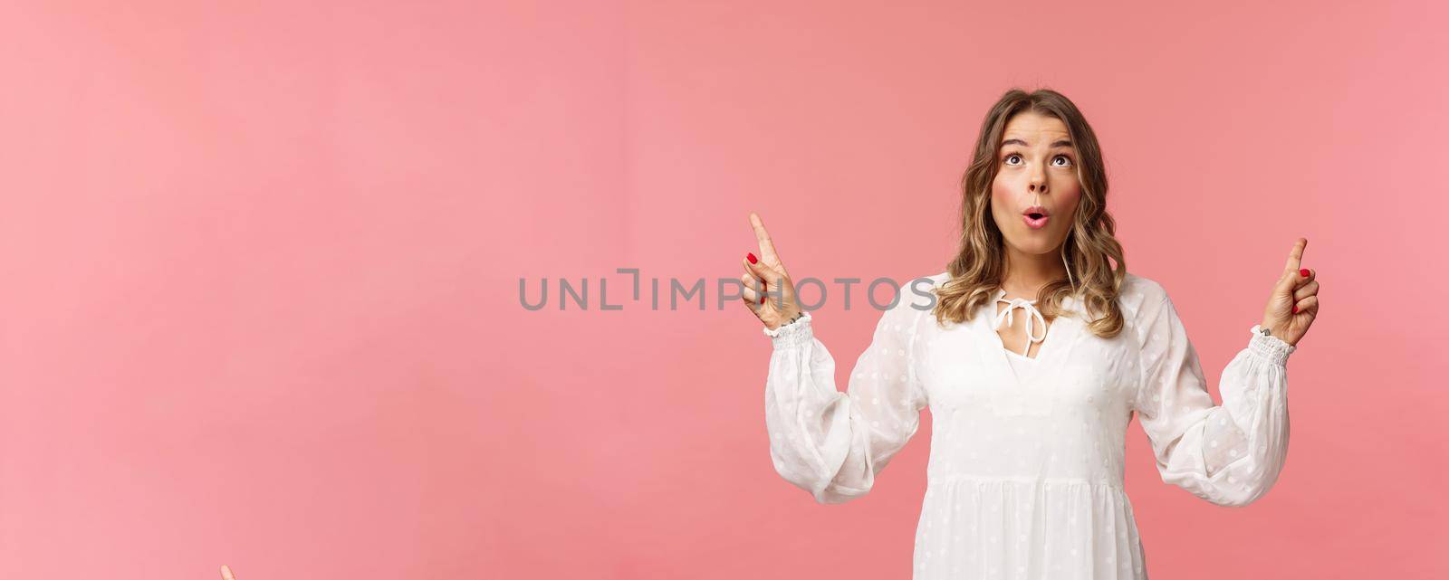 Excited beautiful caucasian blond woman in white dress, fold lips in excitement, looking pointing fingers up at something stunning and breathtaking, enjoying view, pink background.
