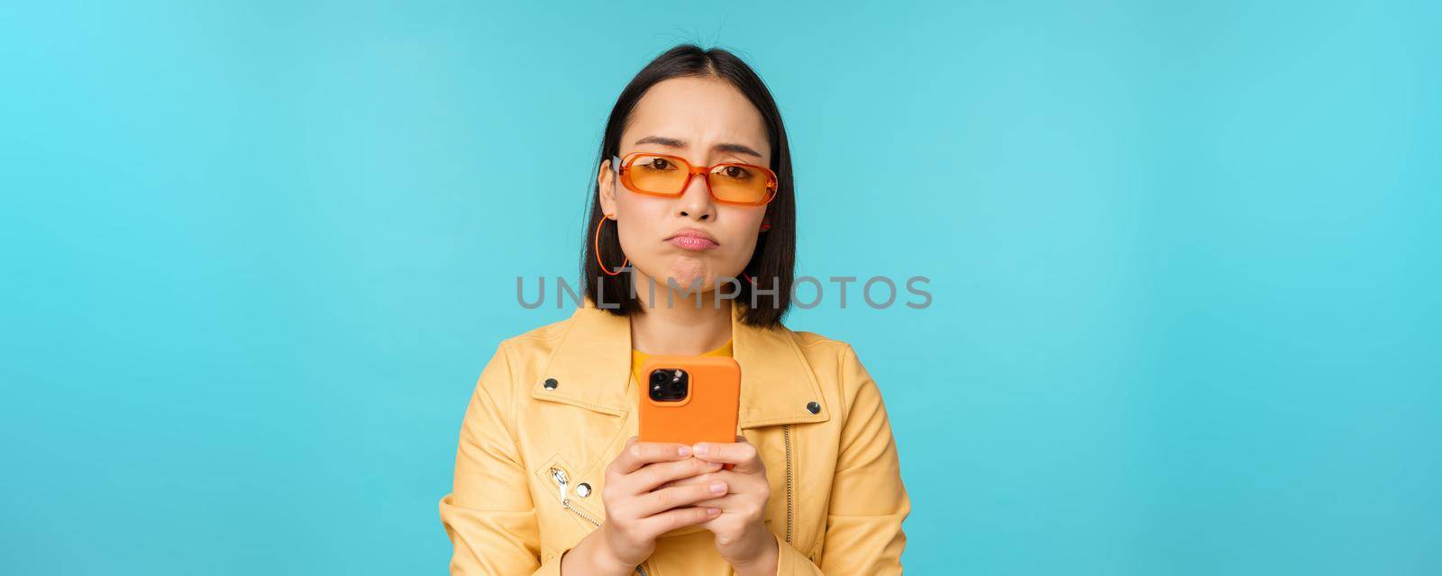 Image of sad asian girl in sunglasses, looking disappointed, holding mobile phone, standing over blue background.