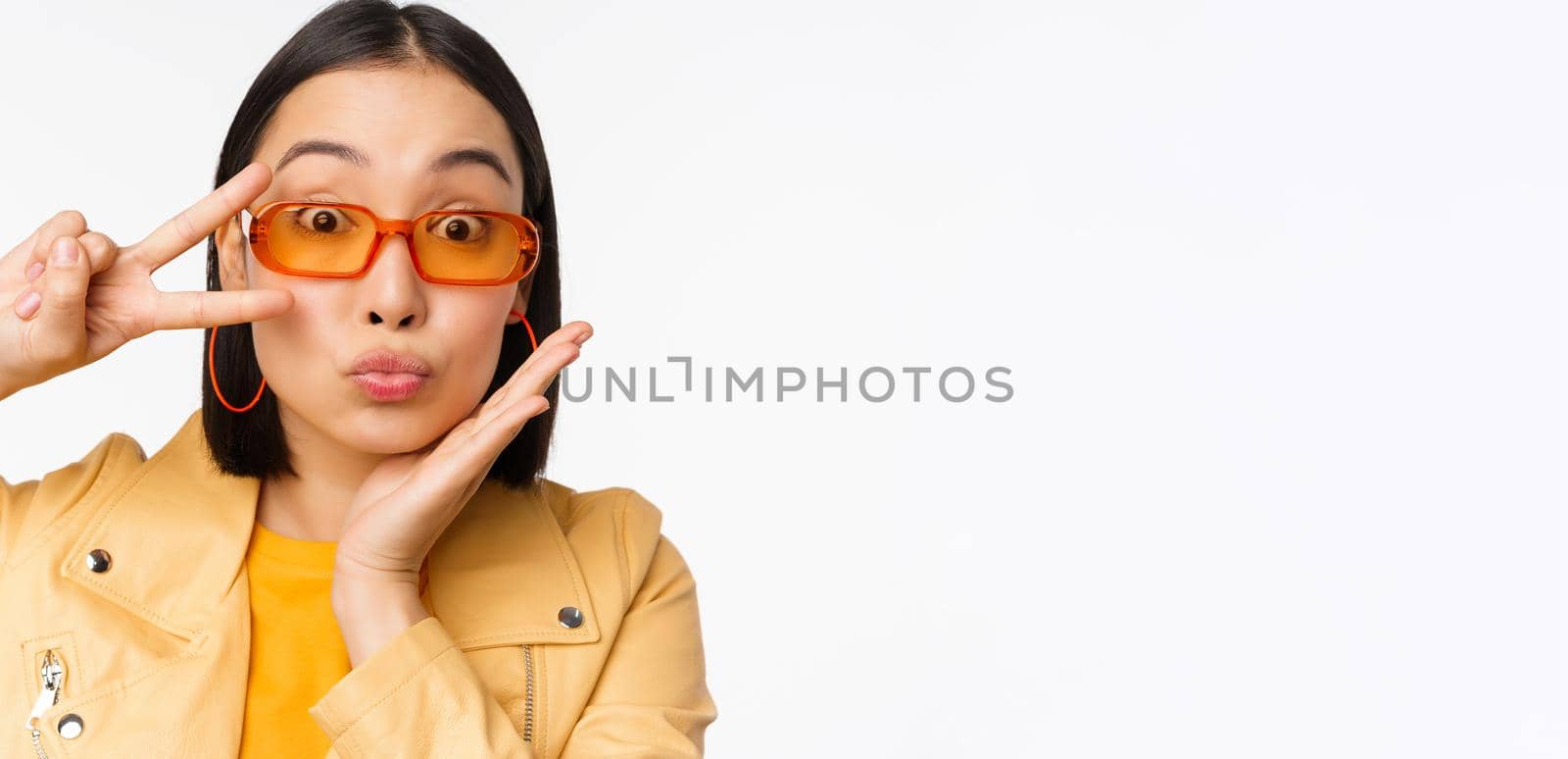 Close up portrait of young asian woman, stylish girl in sunglasses, showing peace, v-sign and smiling, standing over white studio background.