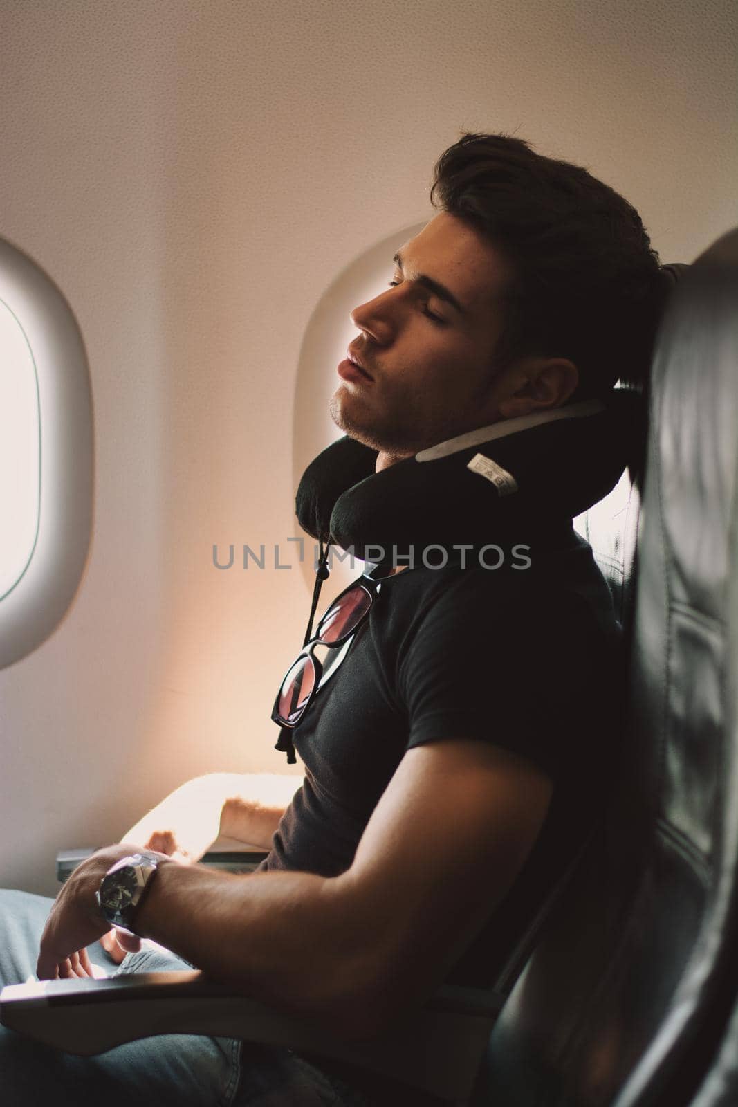 Side view of handsome young man against plane window sleeping with neck pillow