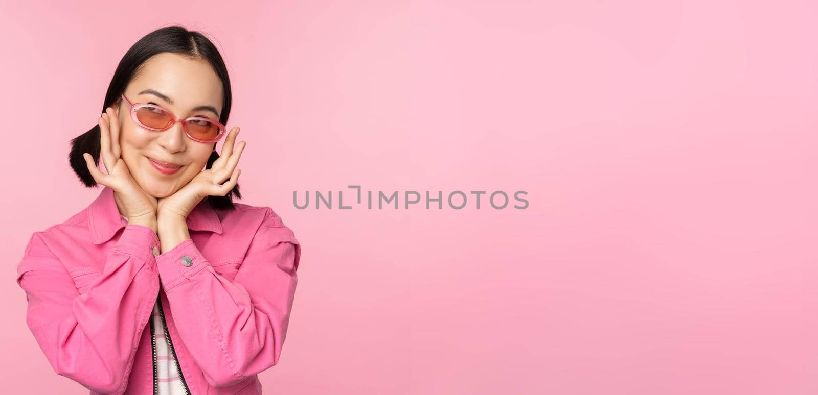 Portrait of stylish cute asian girl, smiling and touching face, looking up dreamy, thoughtful look, standing over pink background.