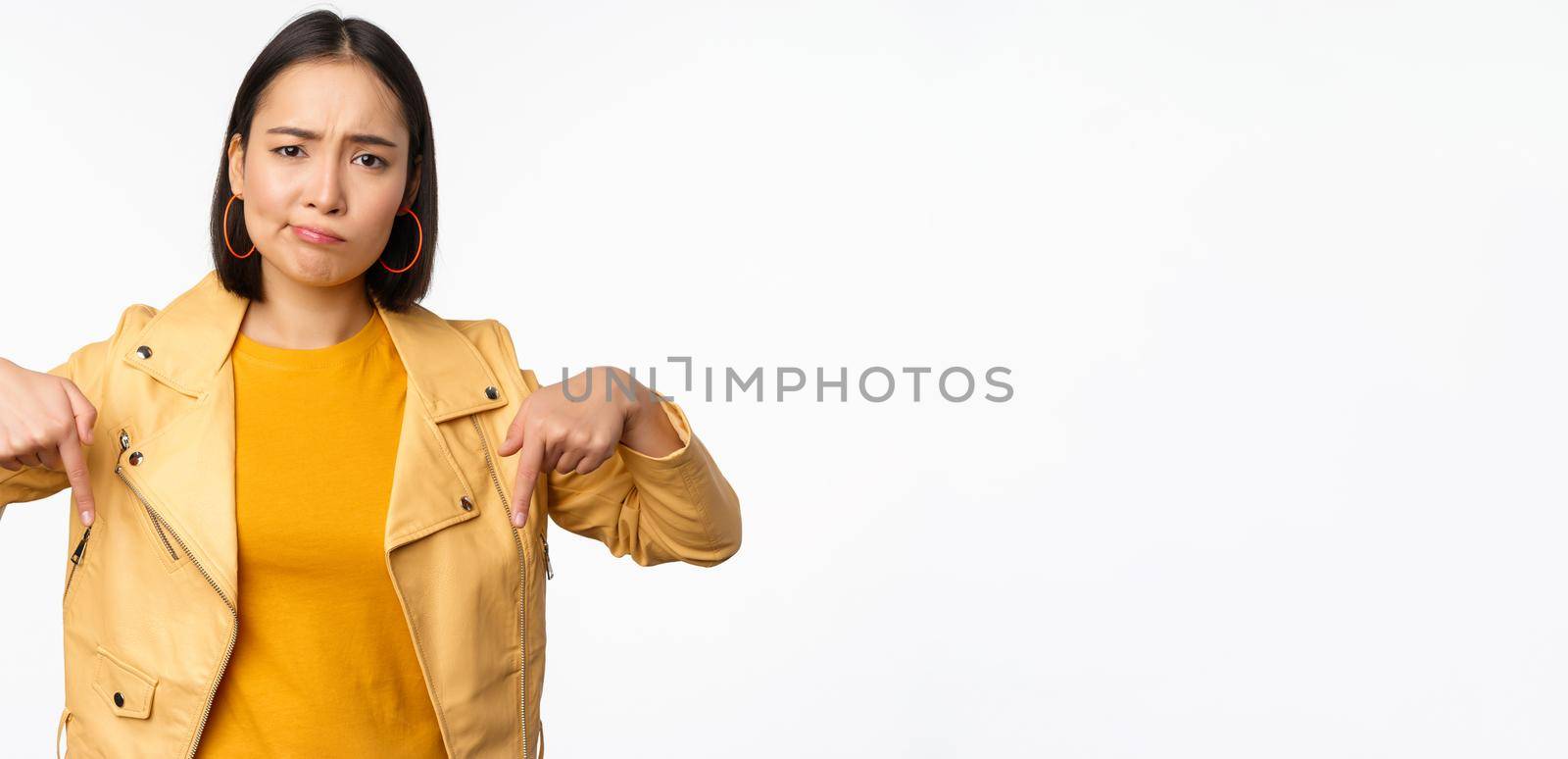 Portrait of disappointed asian woman pointing fingers down, grimacing and showing smth bad, standing over white background. Copy space