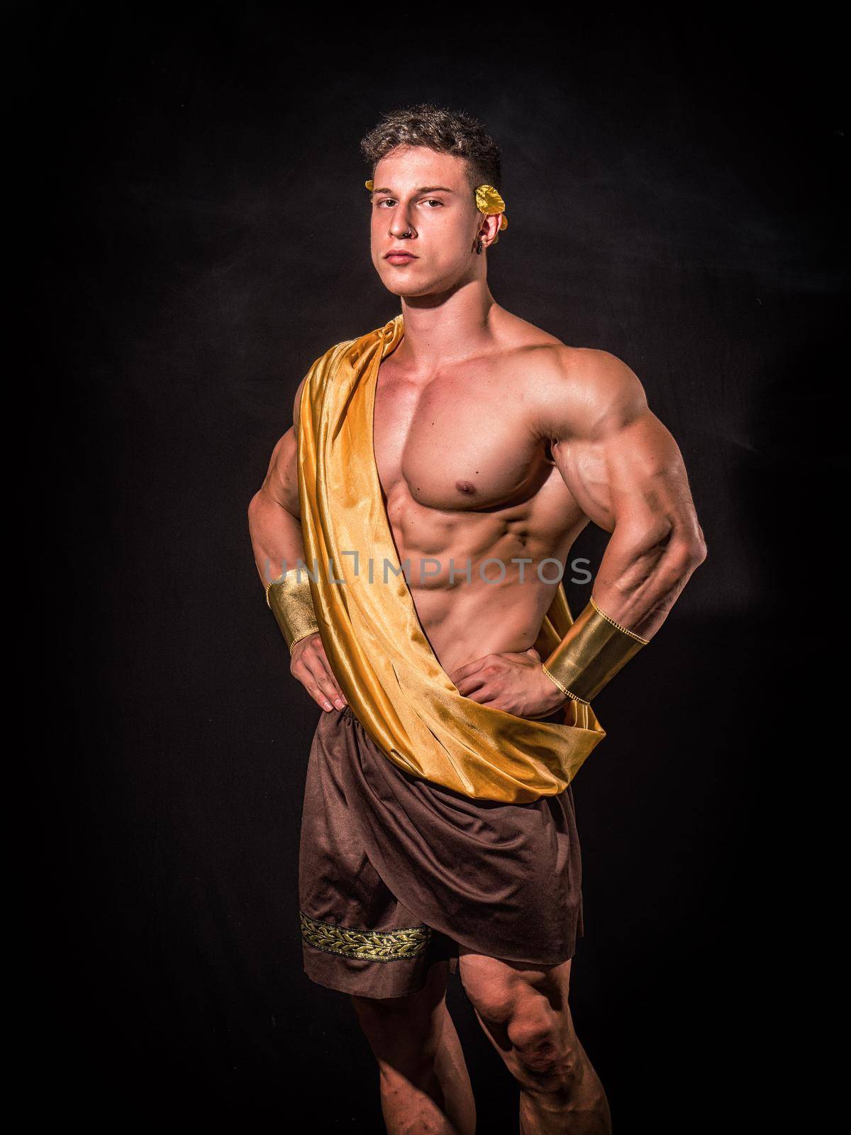 Serious young man in Greek god or Roman aristocrat costume with muscular body, looking at camera on black background