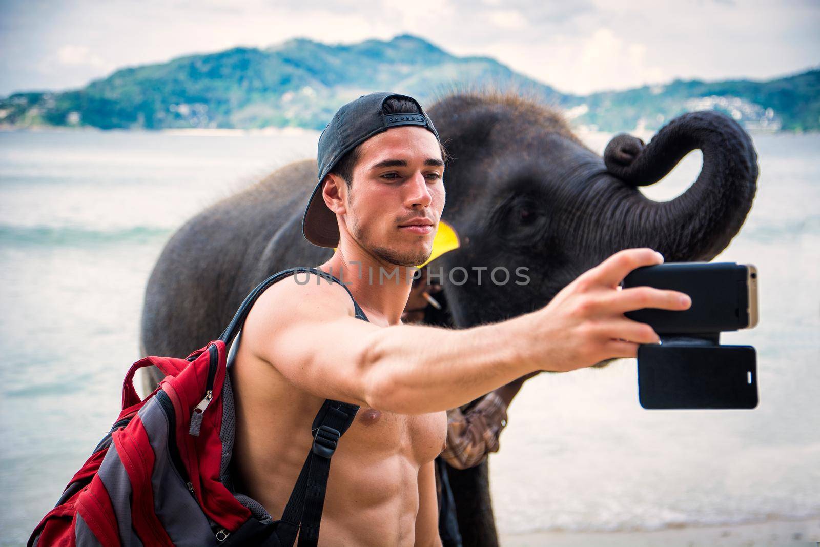 Confident young muscular Hispanic male traveler with naked torso on cap and backpack, taking selfie with elephant on mobile phone, standing on ocean beach during trip in tropical country