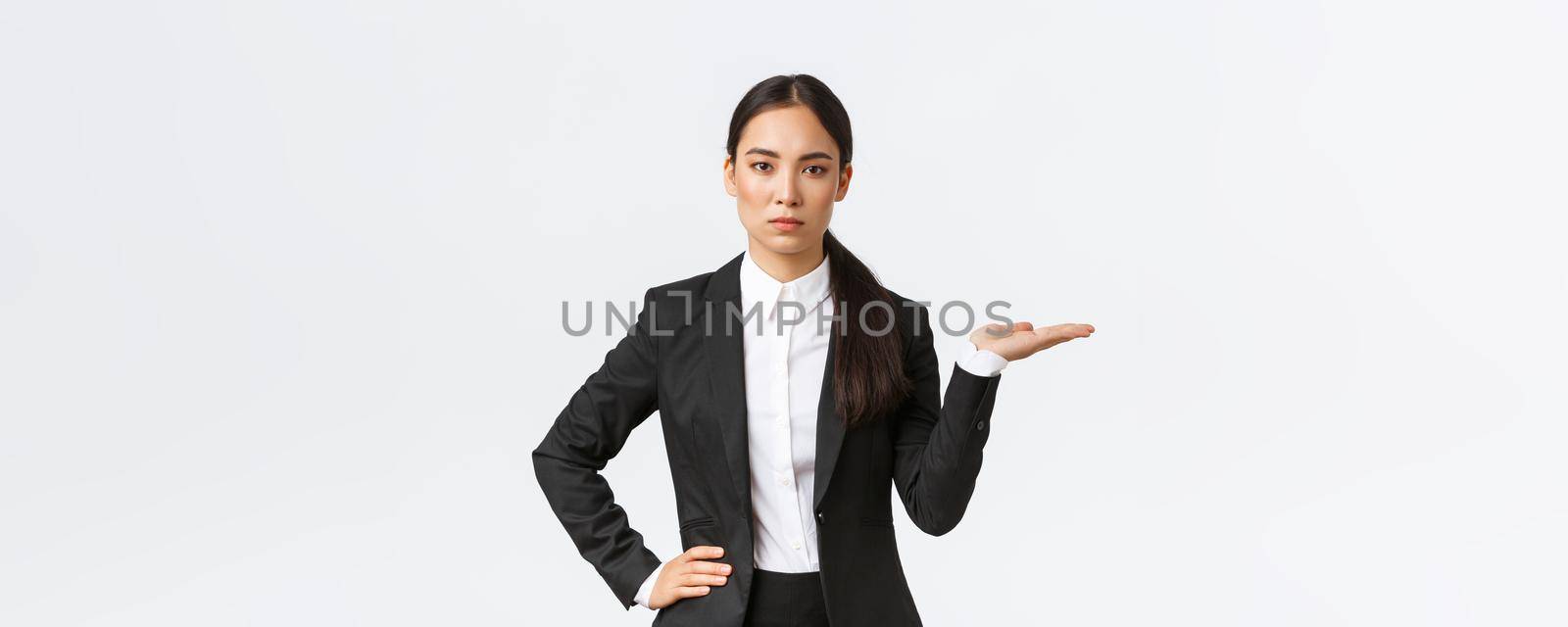 Serious-looking determined young asian businesswoman introduce her project, showing something on palm, holding hand right on blank space, as introduce product, standing in black suit white background.