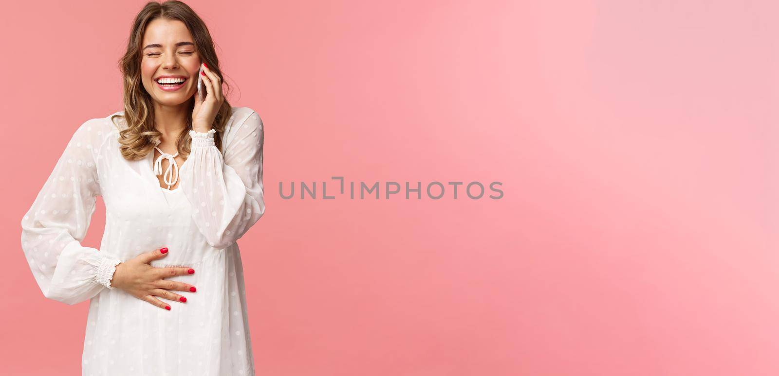 Portrait of joyful happy bright young girl in white dress, having fun, speaking to friend, calling person mobile phone, laughing and touching belly, close eyes smiling, hold smartphone near ear.