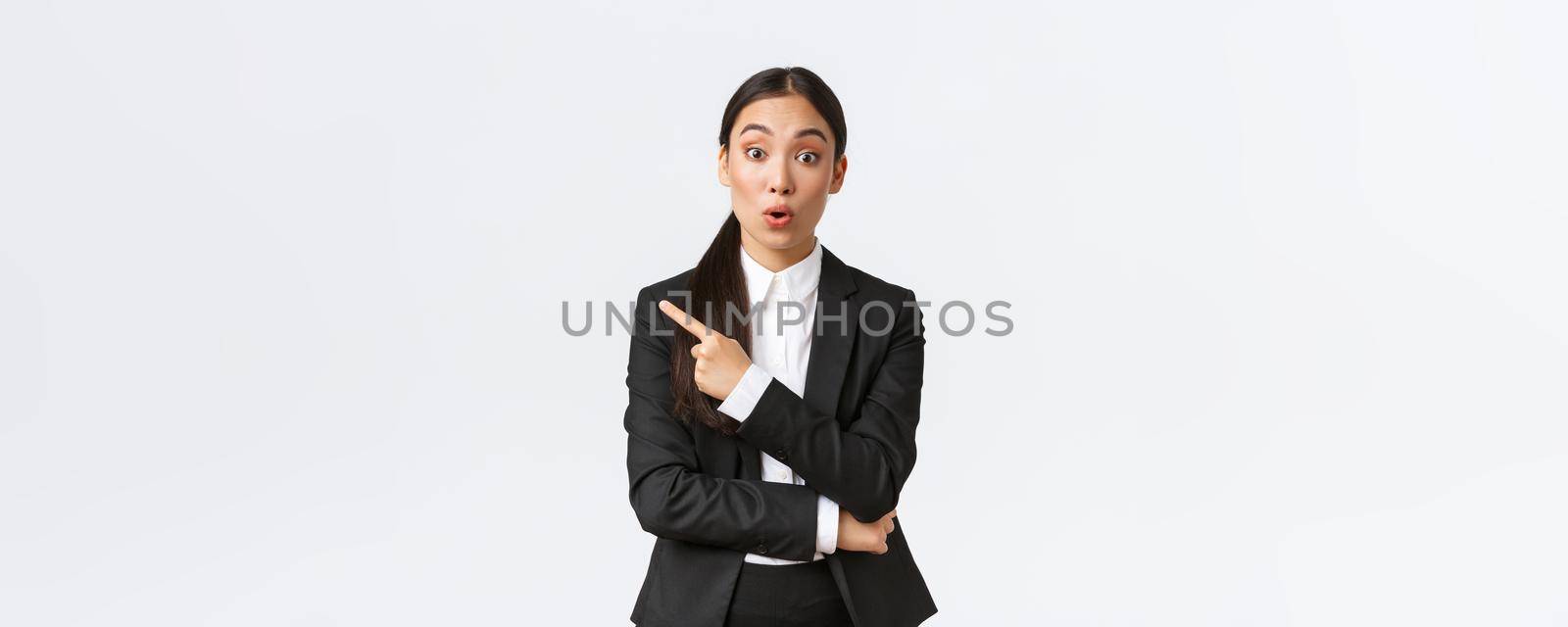 Surprised and intrigued asian female entrepreneur asking question about product, standing interested, pointing finger left to find out details, discussing product or offer with office team.