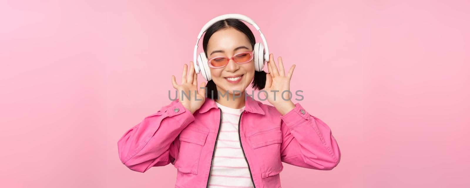 Dancing stylish asian girl listening music in headphones, posing against pink background by Benzoix