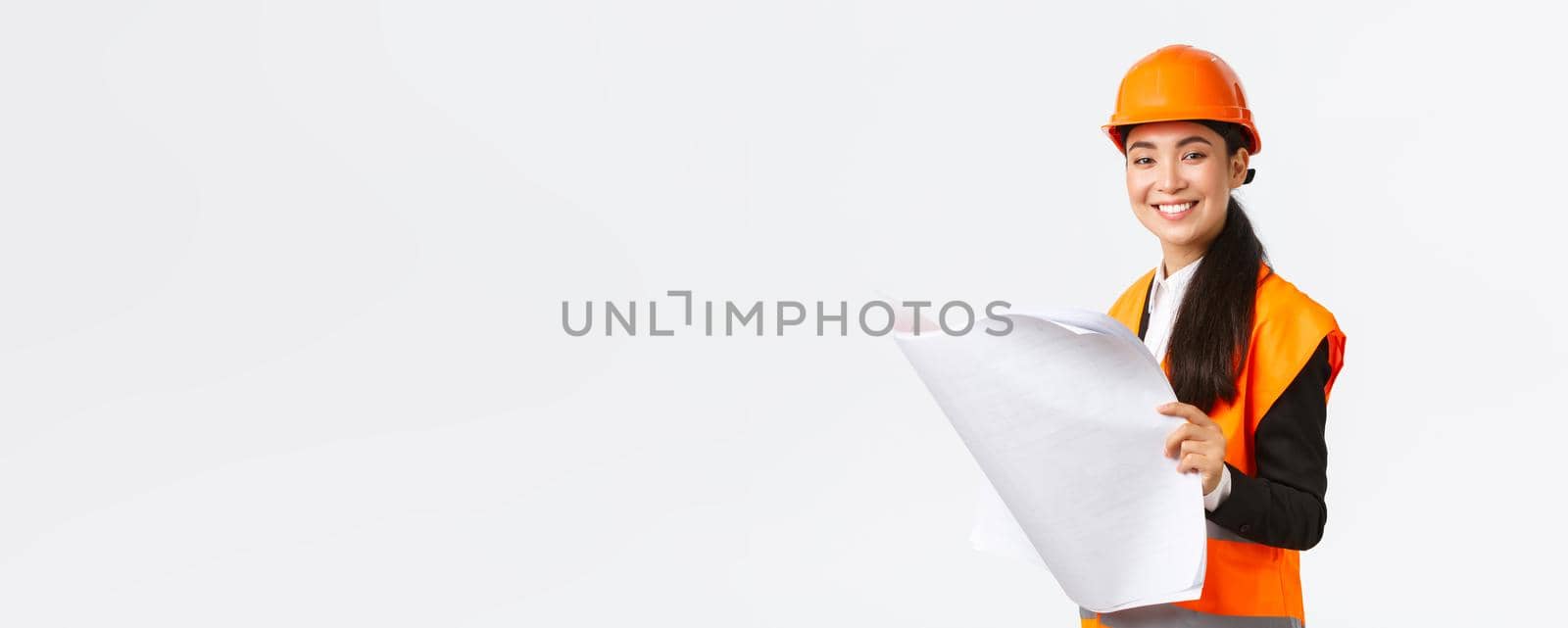 Pleased smiling female asian construction manager following blueprints during building house, looking satisfied at camera, studying architect plan, standing white background in safety helmet by Benzoix